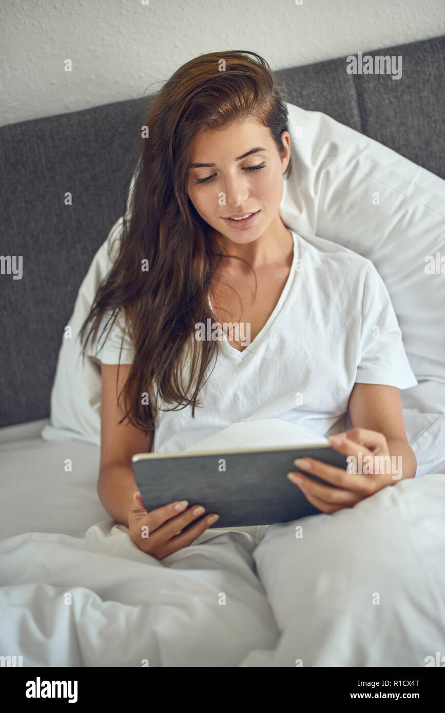 Junge hübsche Frau mit langen brünetten Haare im Bett liegend Unter weißen Decke mit Tablet-PC in ihren Händen, Home Shopping mit Kopie Raum Stockfoto