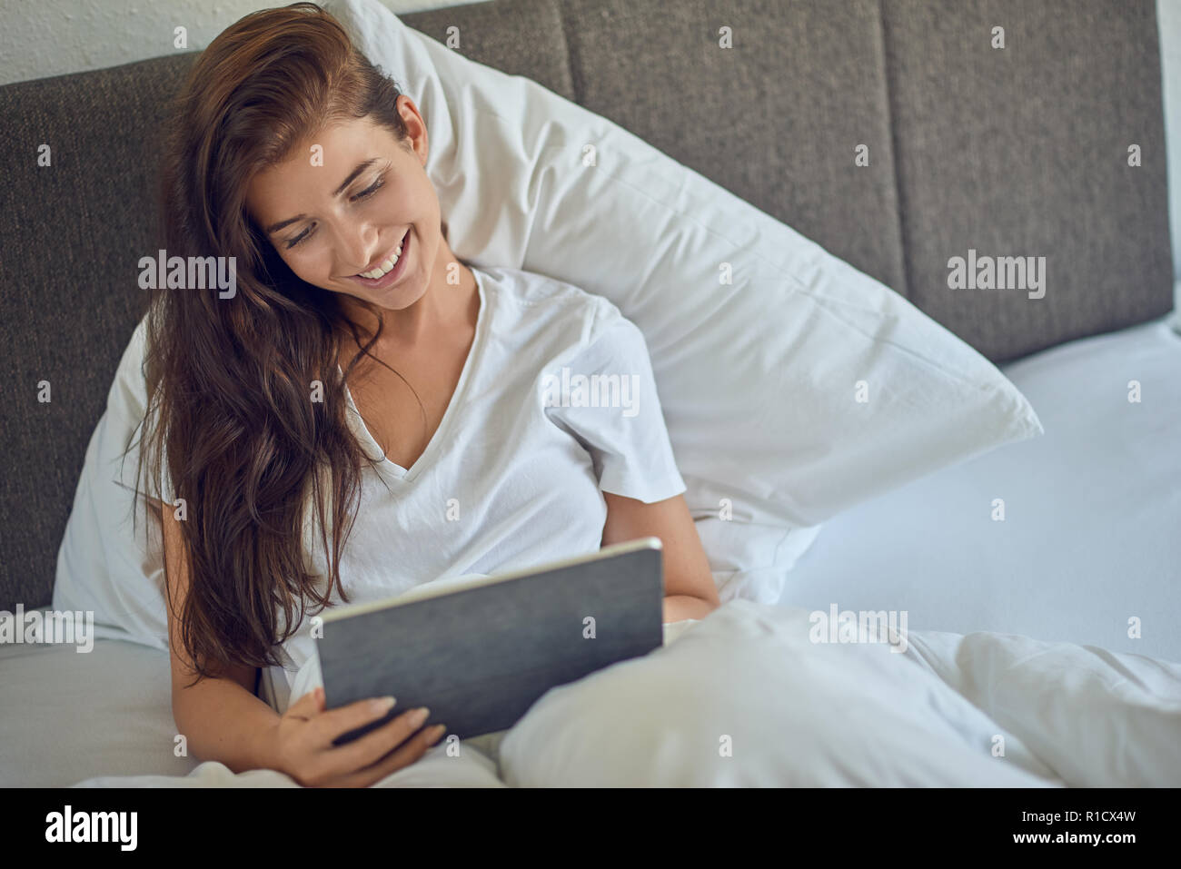 Junge hübsche Frau mit langen brünetten Haare im Bett liegend Unter weißen Decke mit Tablet-PC in ihren Händen, auf Kamera und Lachen mit Kopie spac Stockfoto