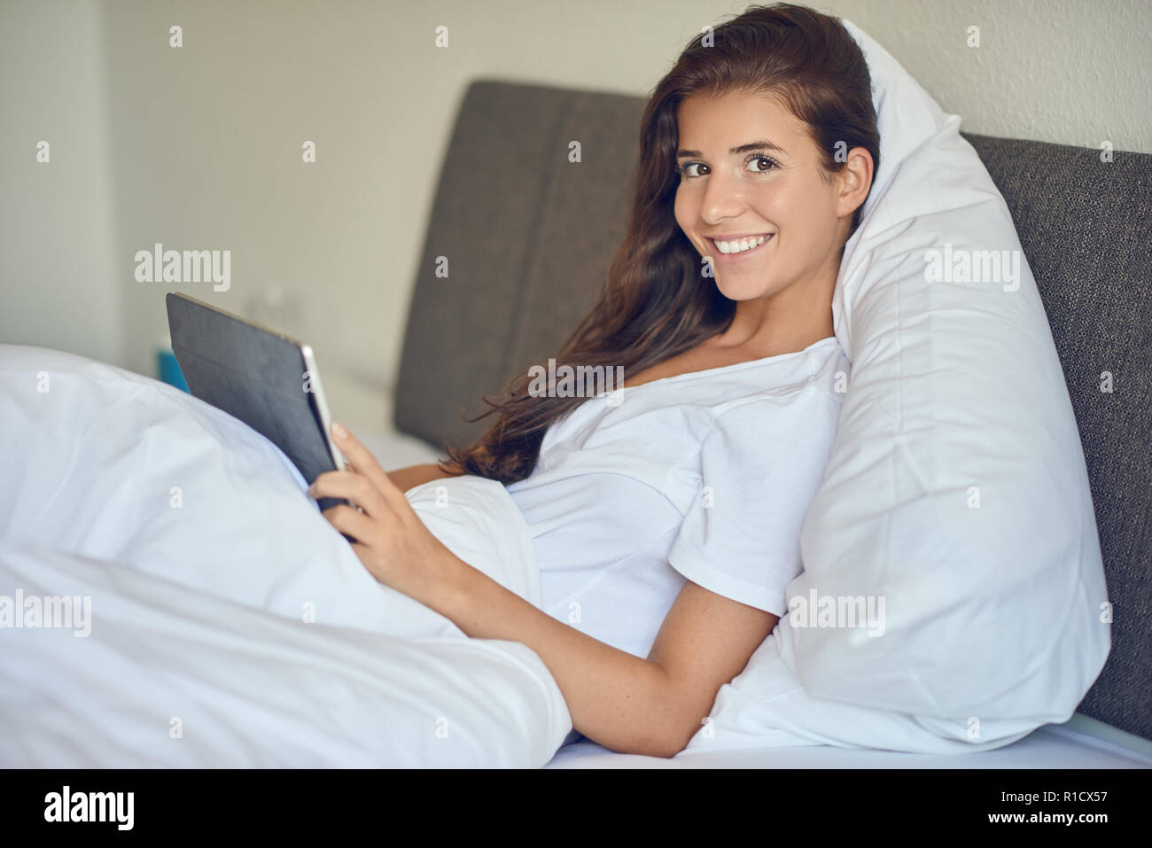 Junge hübsche Frau mit langen brünetten Haare im Bett liegend Unter weißen Decke mit Tablet-PC in ihren Händen, auf Kamera und Lachen mit Kopie spac Stockfoto