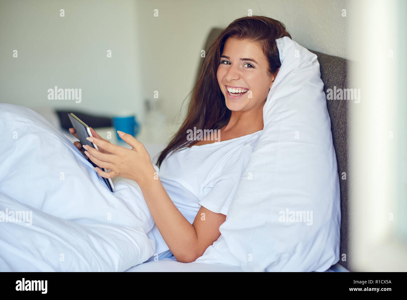 Junge hübsche Frau mit langen brünetten Haare im Bett liegend Unter weißen Decke mit Tablet-PC in ihren Händen, auf Kamera und Lachen mit Kopie spac Stockfoto