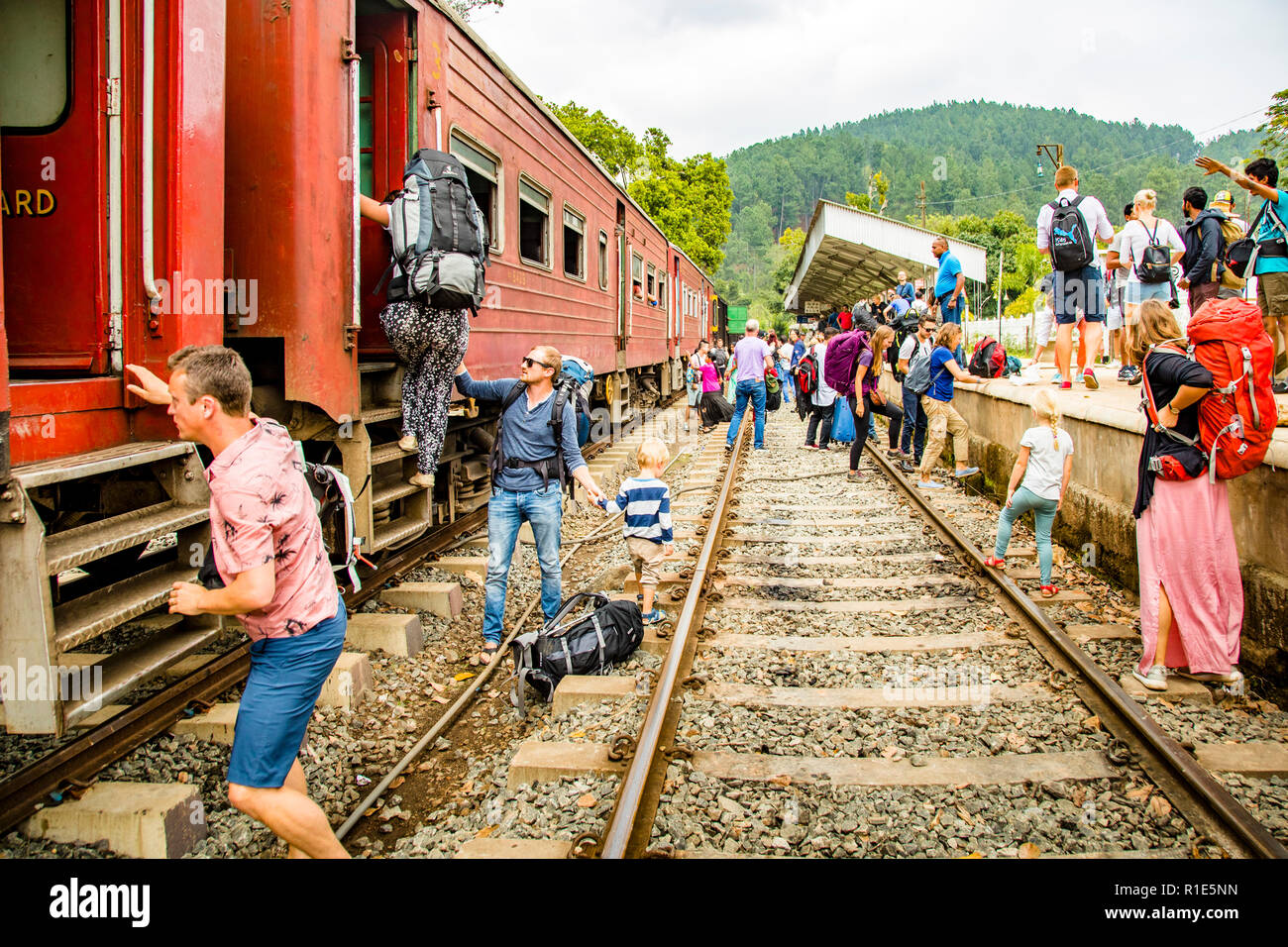 Passagiere steigen ein und überqueren die Gleise in einem Gedränge von Rucksäcken am Bahnhof Ella, Sri Lanka, nachdem der Zug auf dem falschen Bahnsteig eingefahren ist – die Weichen waren wahrscheinlich falsch gestellt. Ella markiert das Ende der malerischen, etwa 65-Kilometer-Linie von Nanu Oya, die 1876 von der britischen Kolonialverwaltung erbaut wurde, um Tee, Zimt, Pfeffer und Muskatnuss aus den Hochländern in die Hafenstadt Colombo zu transportieren. Stockfoto