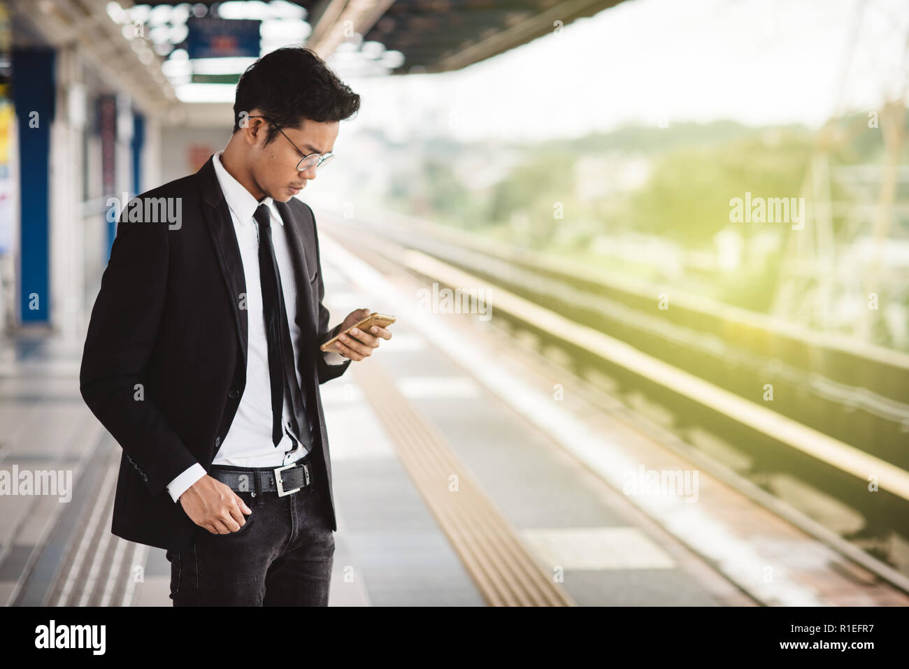 Jungen asiatischen hipster Geschäftsmann mit Smartphone warten am Bahnsteig. Konzept der drahtlosen Technologie, den öffentlichen Verkehrsmitteln und Business Travel Stockfoto