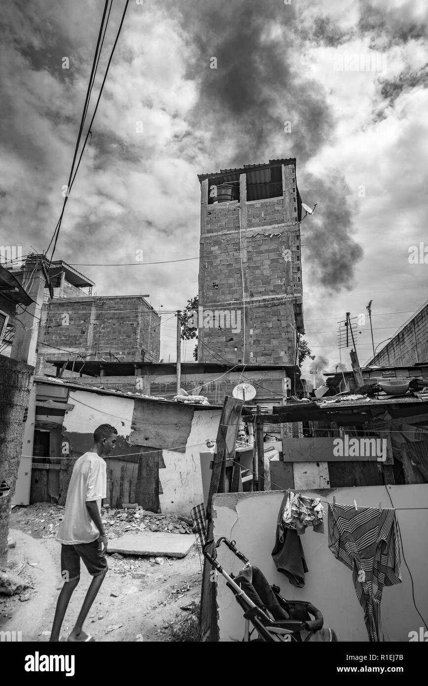 Schwarzer Mann zu Fuß durch den Slum von Rio de Janeiro gebaut von einfachen Hütten mit schwarzem Rauch in den Himmel, die bei der Verbrennung von Müll Stockfoto