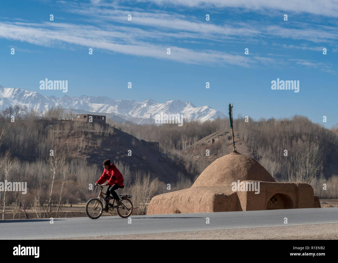Junge Radfahren auf Asphalt in die Provinz Bamyan Valley mit einem Schrein und Snow-Capped Bergen im Hintergrund, Provinz Bamyan, Afghanistan Stockfoto