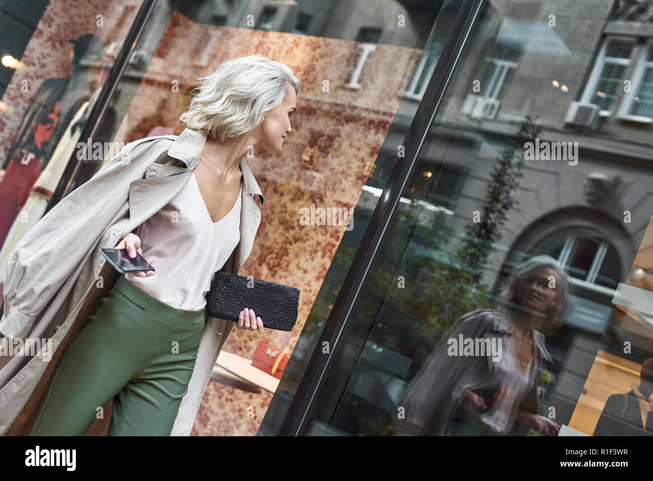 Mode. Junge stilvolle Frau, die zu Fuß auf der Straße halten Sie das Smartphone auf Reflexion im Fenster suchen Stockfoto