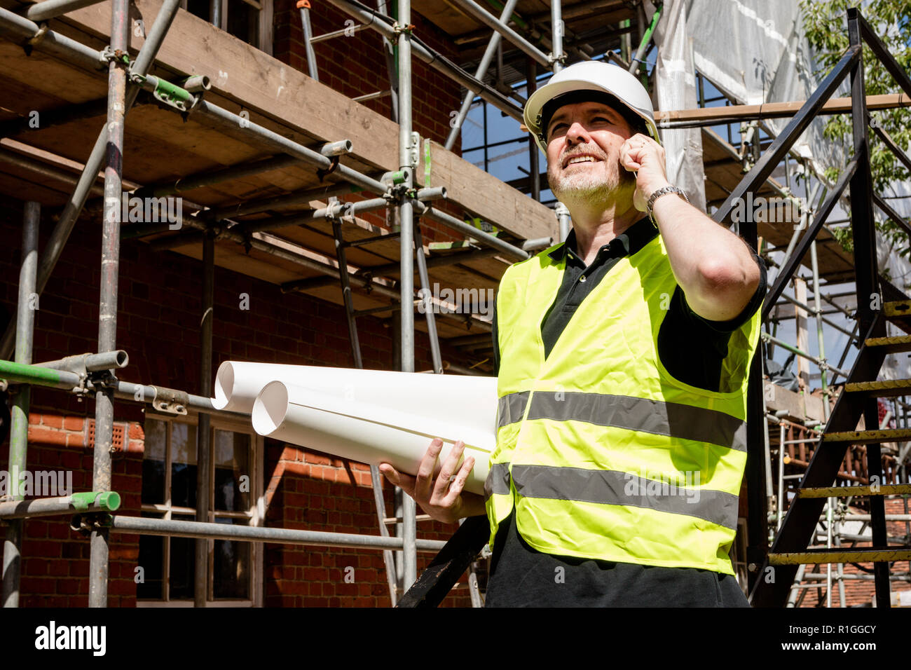 Männliche Bauarbeiter, Polier oder Architekt auf der Baustelle über sein Mobiltelefon und Holding gerollt Baupläne Stockfoto