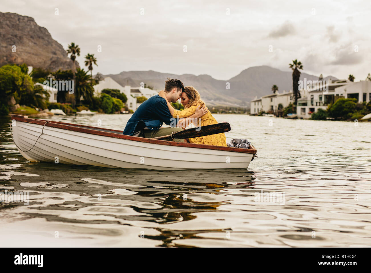 Junges Paar in einem Boot die Köpfe gegenseitig Holding berühren sitzen. Paar auf einem Boot Datum in einem See mit Hügeln und Häuser im Hintergrund. Stockfoto