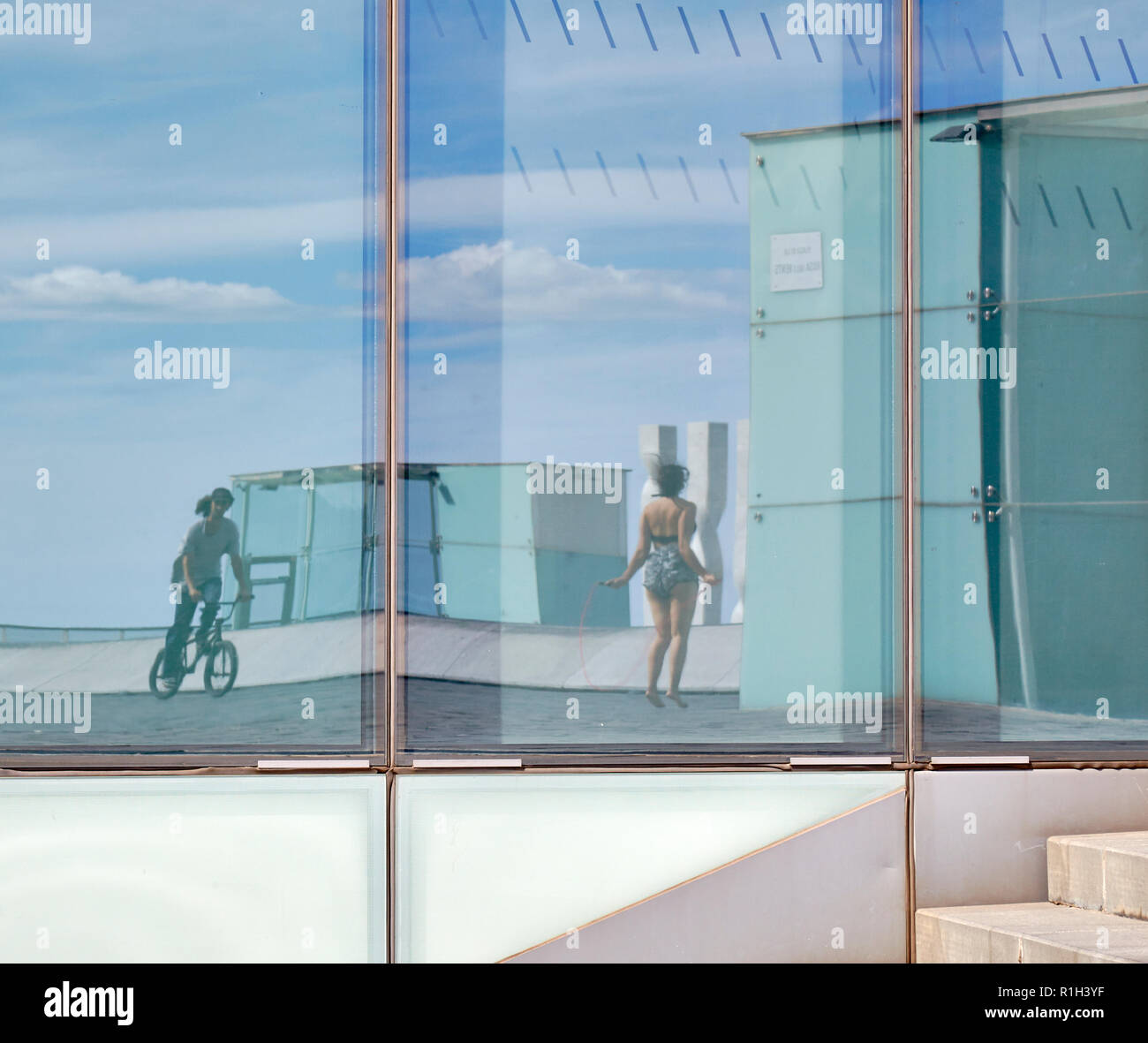 Reflexionen über eine Glasfassade. Ein Kind mit einem Fahrrad und einem Mädchen Rope Skipping. Stockfoto