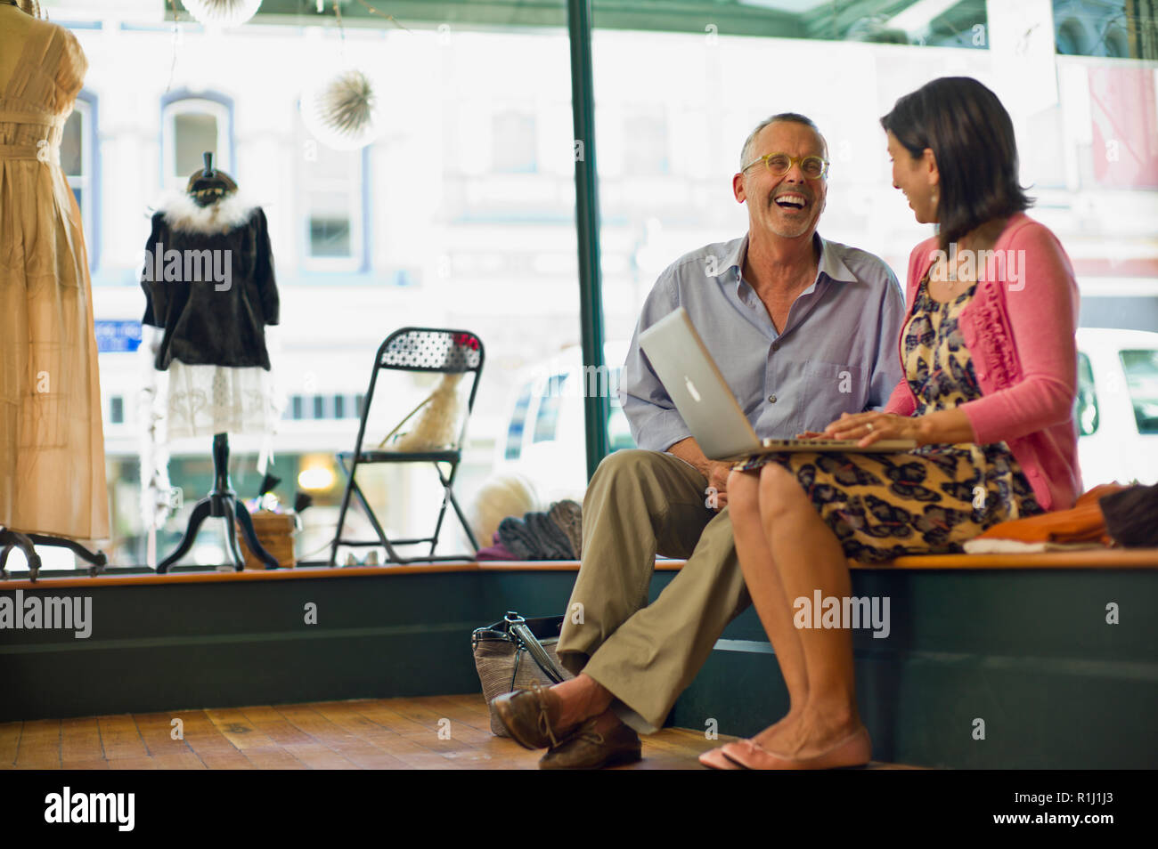Lachend reifer Mann neben ihr sitzt Frau, als sie ihr Laptop in einem Bekleidungsgeschäft verwendet. Stockfoto