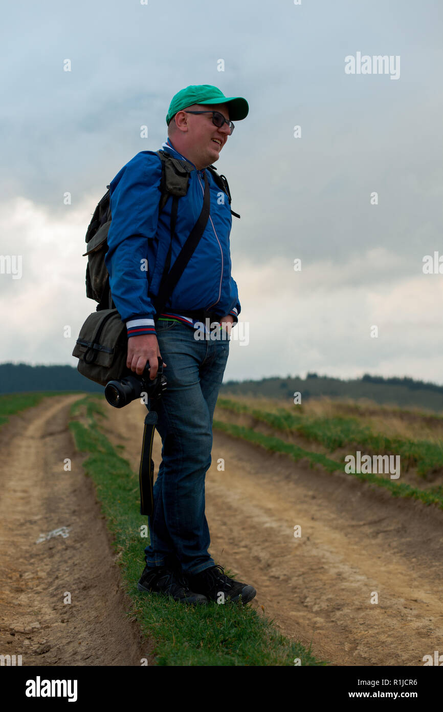 Fotograf in blau Windjacke und Jeans mit einer Kamera und einem Rucksack steht auf einem Feldweg in den Bergen Stockfoto