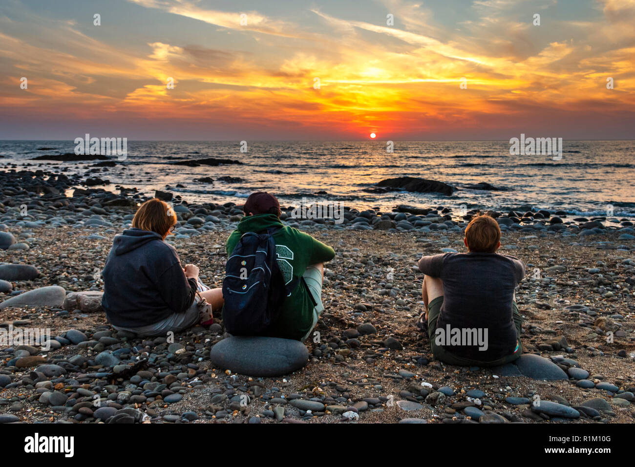 Dreiköpfige Familie (Mutter, Sohn, Tochter), die an einem steinigen Strand sitzt und den Sonnenuntergang in Westward Ho!, Devon, Großbritannien, beobachtet Stockfoto