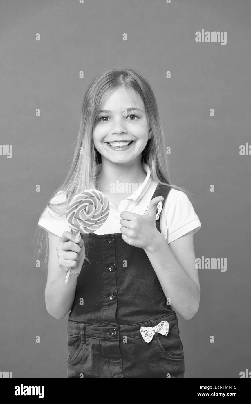 Mädchen mit bunten Lutscher. Süße Kindheit Konzept. Kind mit langen Haaren gerne Süßigkeiten. Mädchen mit lächelnden Gesicht hält Giant candy in der Hand, violett unterlegt. Kind in rosa Overalls, Daumen hoch. Stockfoto