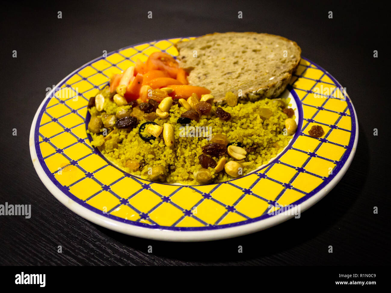 Futternapf auf schwarzem Holz- Hintergrund. Couscous, Tomaten, Erdnüsse, Rosinen, Brot. Gelb Blau Platte. Nahaufnahme. Stockfoto