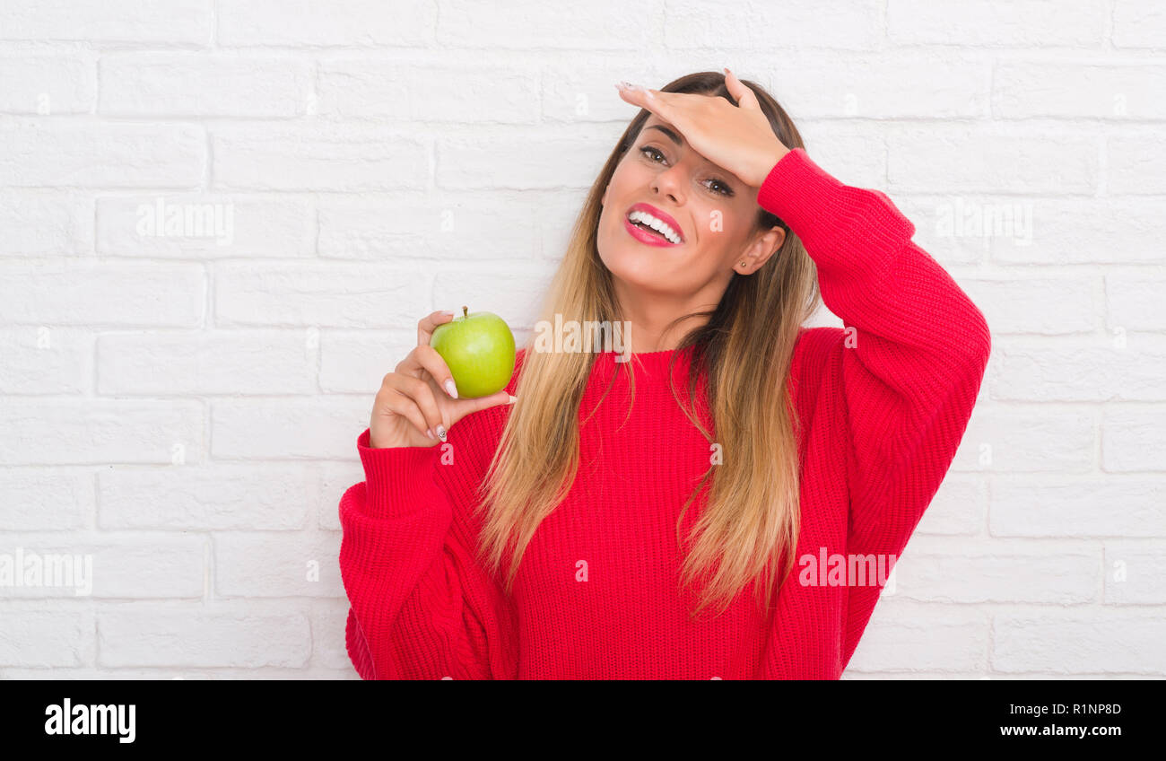 Junge erwachsene Frau über weiße Mauer essen frischen grünen Apfel mit Hand betonte am Kopf, mit Scham und Überraschung Gesicht schockiert, wütend und frustriert Stockfoto