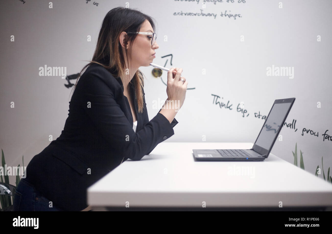 Eine junge Frau denken, 30-39 Jahre alt, Seitenansicht, obere Körper geschossen. Arbeiten am Laptop. Wand mit kreativen Zitate hinter sich. Stockfoto