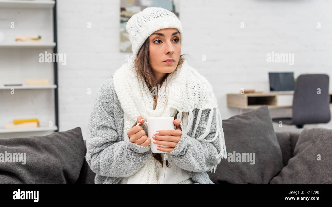 Zugefrorene junge Frau in warme Kleidung holding Tasse wärmenden Tee, während auf der Couch zu Hause sitzen Stockfoto