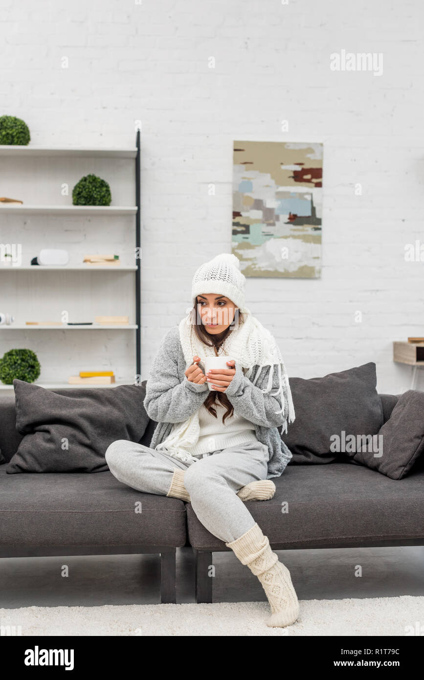 Zugefrorene junge Frau in warme Kleidung Holding heiße Tasse Tee beim Sitzen auf der Couch zu Hause Stockfoto