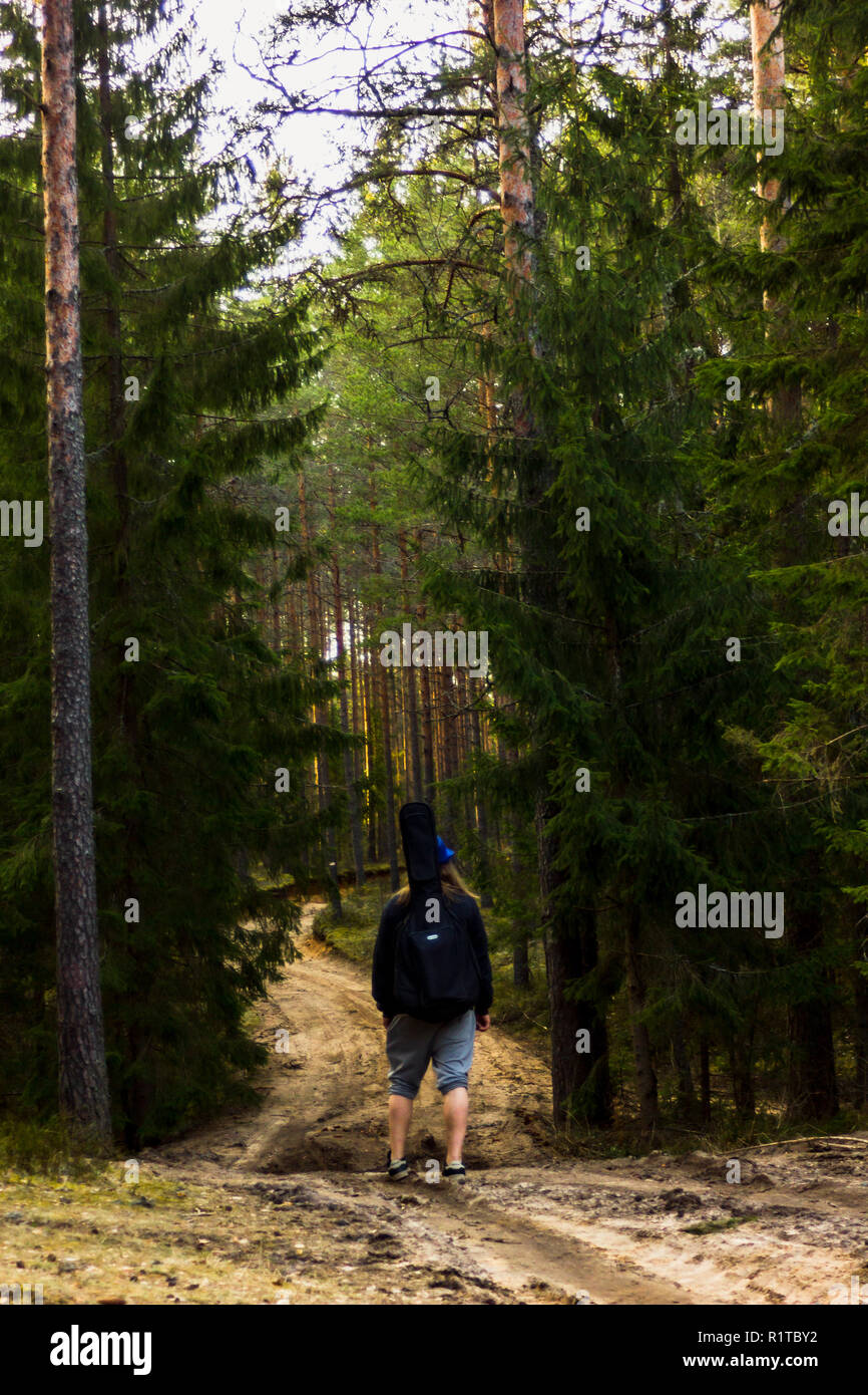 Jung und hübscher Kerl zu Fuß auf der Straße in den Wald mit einer Gitarre auf seinen Schultern und blauen Hut auf dem Kopf auf einem sonnigen Frühling Tag Stockfoto