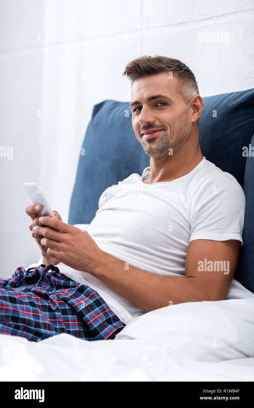 Lächelnd Mann im weißen T-Shirt mit Kamera und Sie ihr Smartphone im Bett zu Hause. Stockfoto
