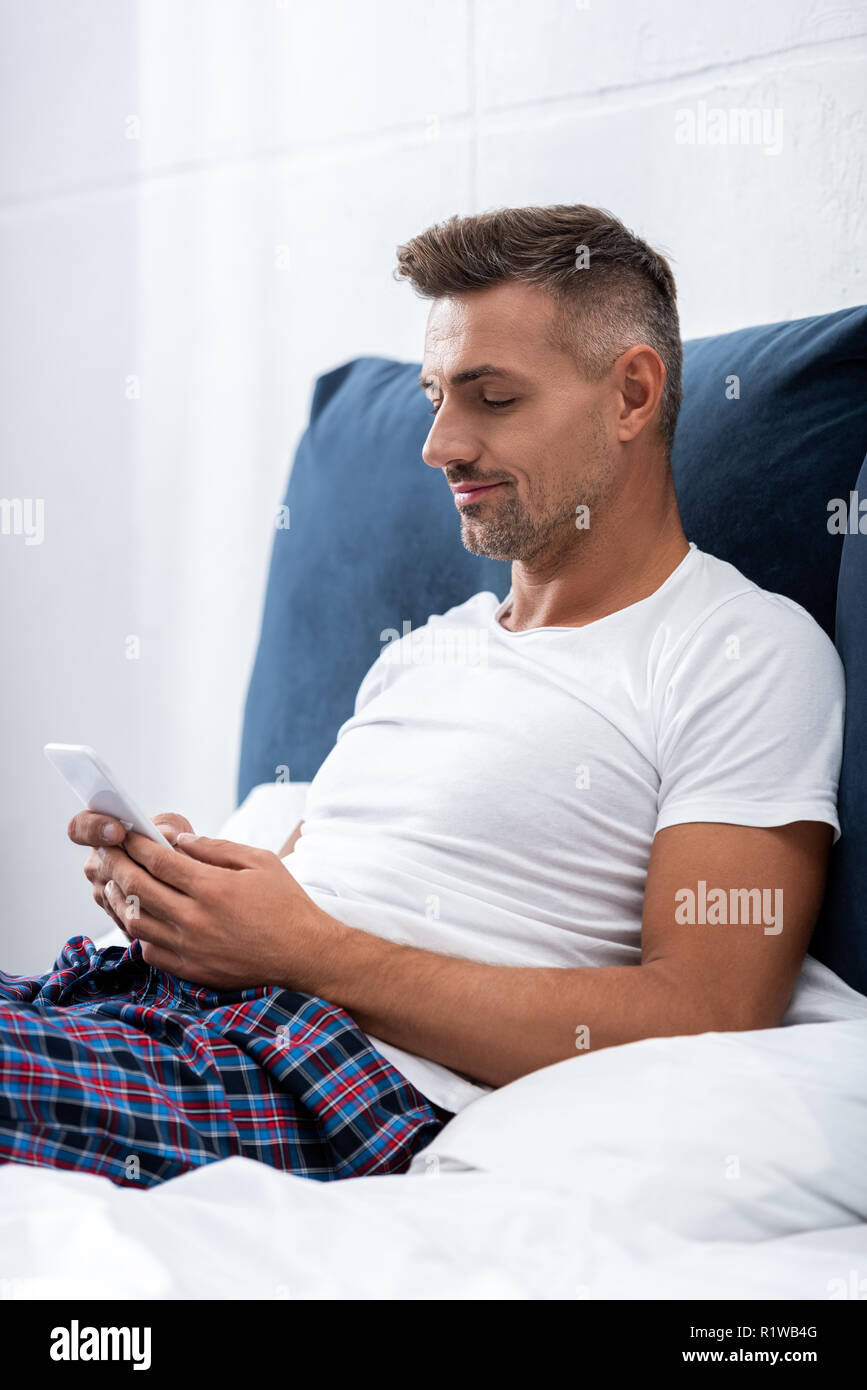 Mann im weißen T-Shirt mit Smartphone während der Zeit im Bett zu Hause. Stockfoto
