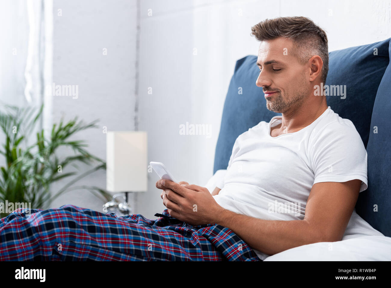 Fröhlicher Mann im weißen T-Shirt mit Smartphone während der Zeit im Bett zu Hause. Stockfoto