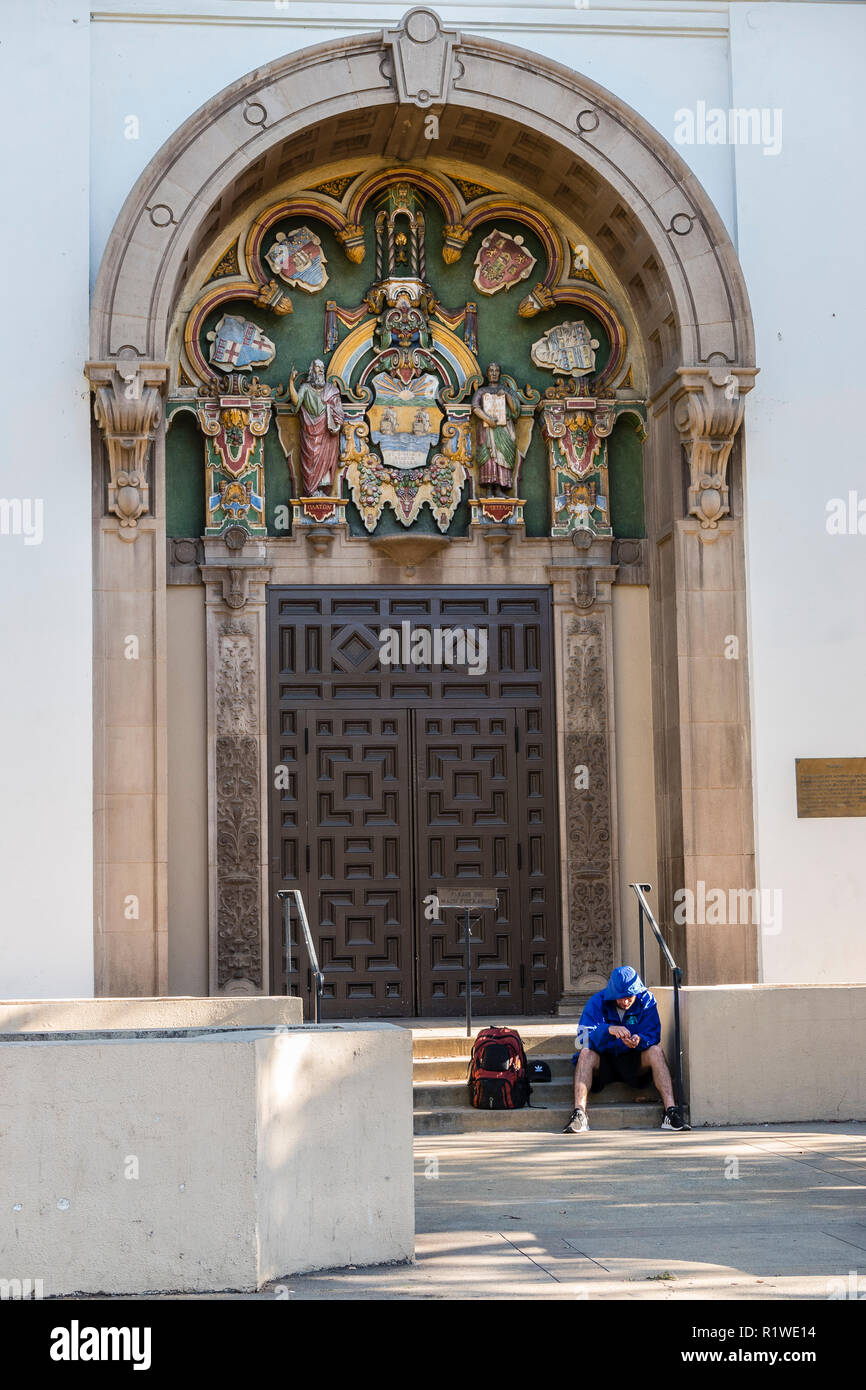 Ein obdachloser Mann sitzt auf den Stufen zur Stadt Bibliothek mit seinem Rucksack und das Tragen eines Poncho und Shorts. Stockfoto