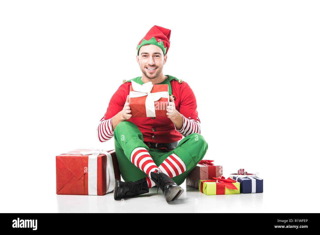 Glücklich lächelnde Mann in Weihnachtself kostüm sitzen in der Nähe von Pile Geschenke und Geschenk isoliert auf weißem Stockfoto