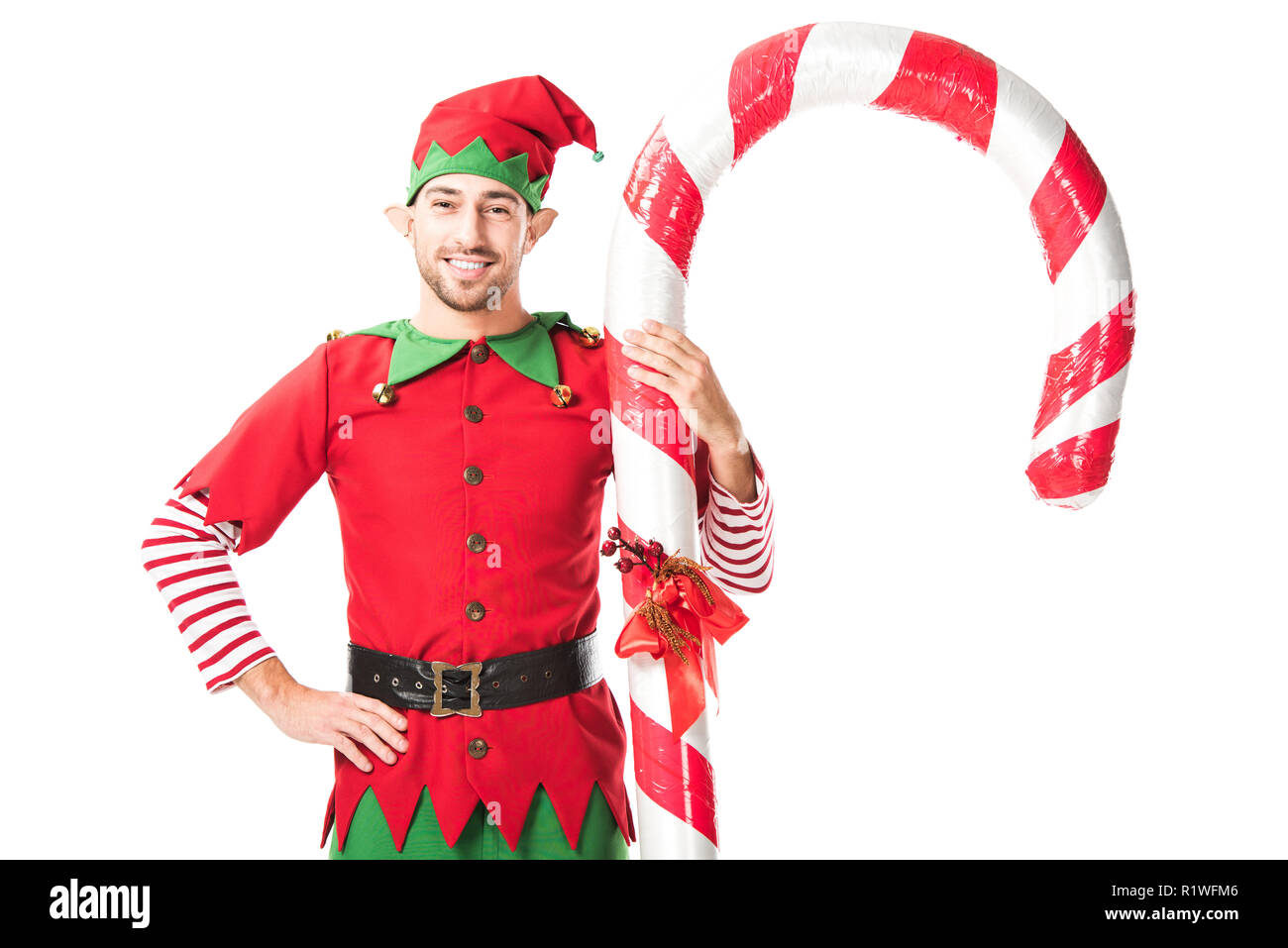 Lächelnd Mann in Weihnachtself kostüm mit der Hand auf den Hüften in der Nähe von Big Candy Cane stehen isoliert auf weißem Stockfoto