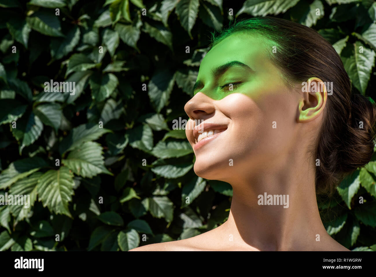 Junge lächelnde Frau mit grünen Schatten auf dem Gesicht und Blätter im Hintergrund Stockfoto