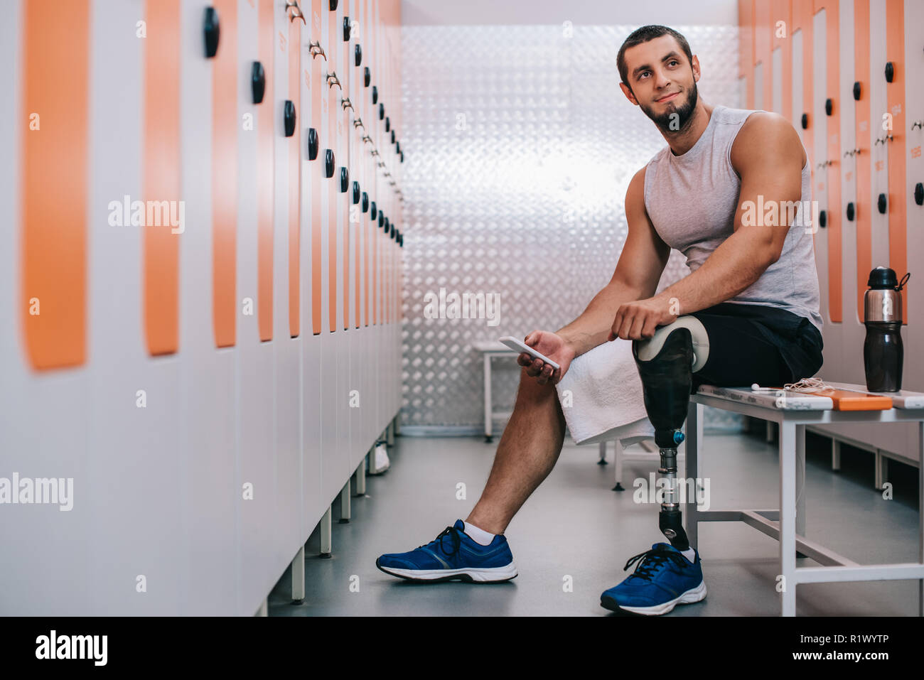 Lächelnden jungen Sportler mit künstlichen Bein sitzt auf der Bank an der Turnhalle Umkleiden und Sie ihr Smartphone Stockfoto