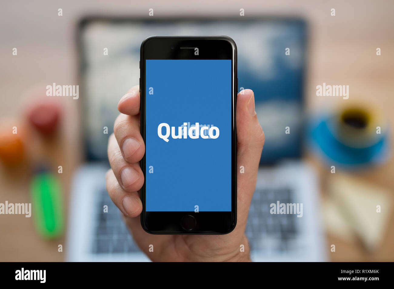 Ein Mann schaut auf seinem iPhone die zeigt die Quidco logo, während saß an seinem Computer Schreibtisch (nur redaktionelle Nutzung). Stockfoto