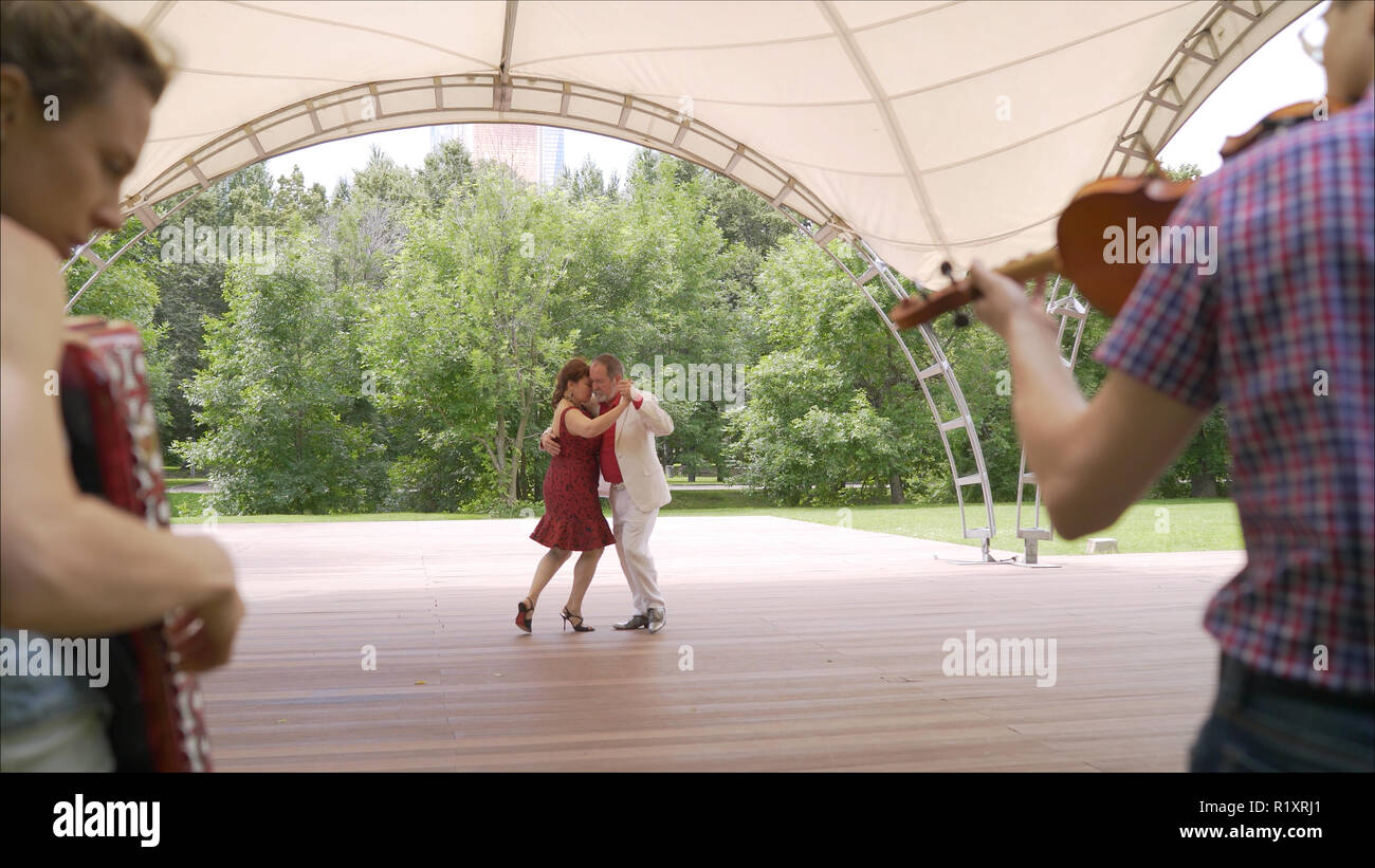 Akkordeon und Geige spielen im Park. Eine Frau in einem roten Kleid und ein Mann in einem weißen Anzug sind Tango tanzen. Ein älteres Paar Tango tanzen im Park. Stockfoto