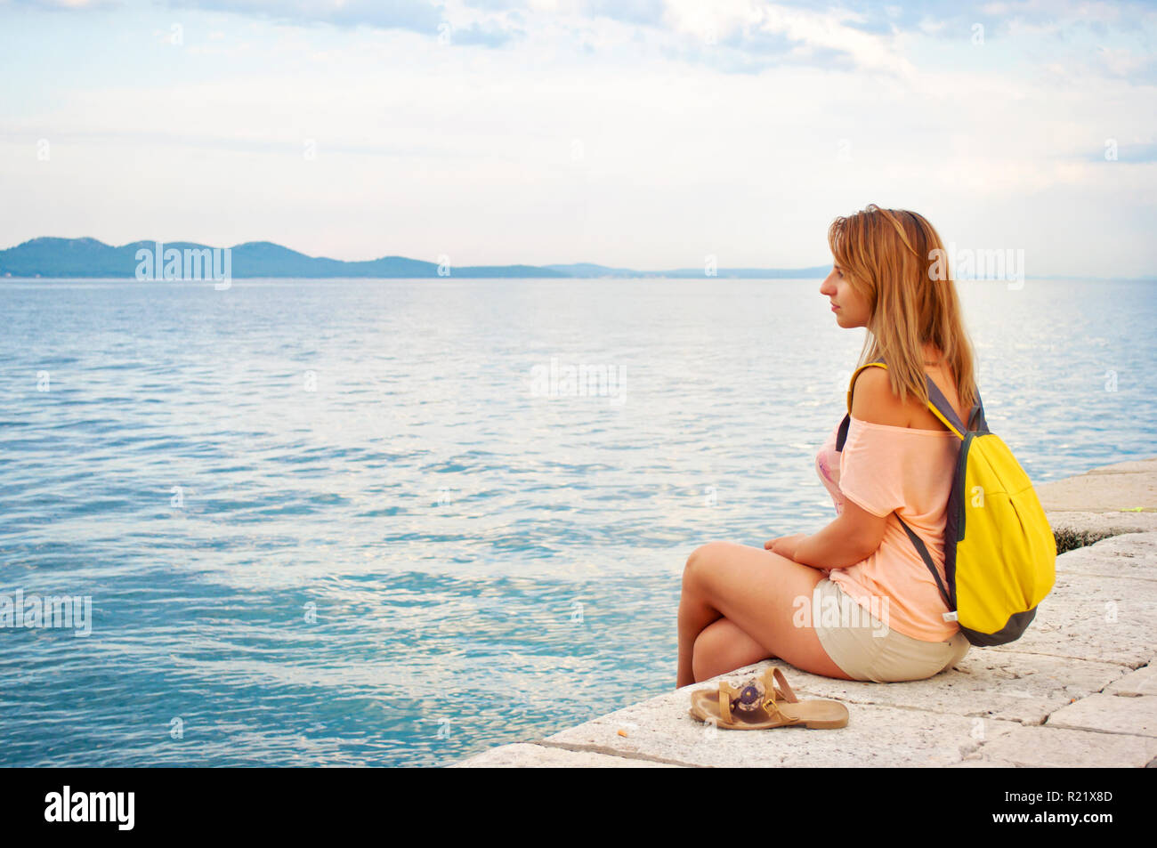 Ein junges Mädchen mit einem Rucksack friedlich seitlich in der Nähe der Küste suchen wehmütig auf die Hügel in der Ferne, die Beine gekreuzt, w Stockfoto