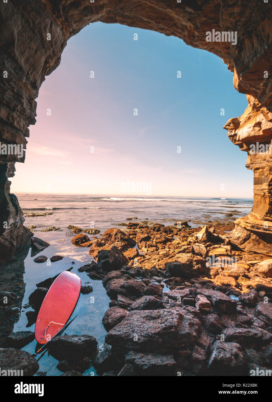 Abondoned rot Surfbrett am Eingang zu einer Höhle. Viele Felsen im Vordergrund und einen Sonnenuntergang Marine im Hintergrund. Entlang Sonnenuntergang Cli entfernt Stockfoto