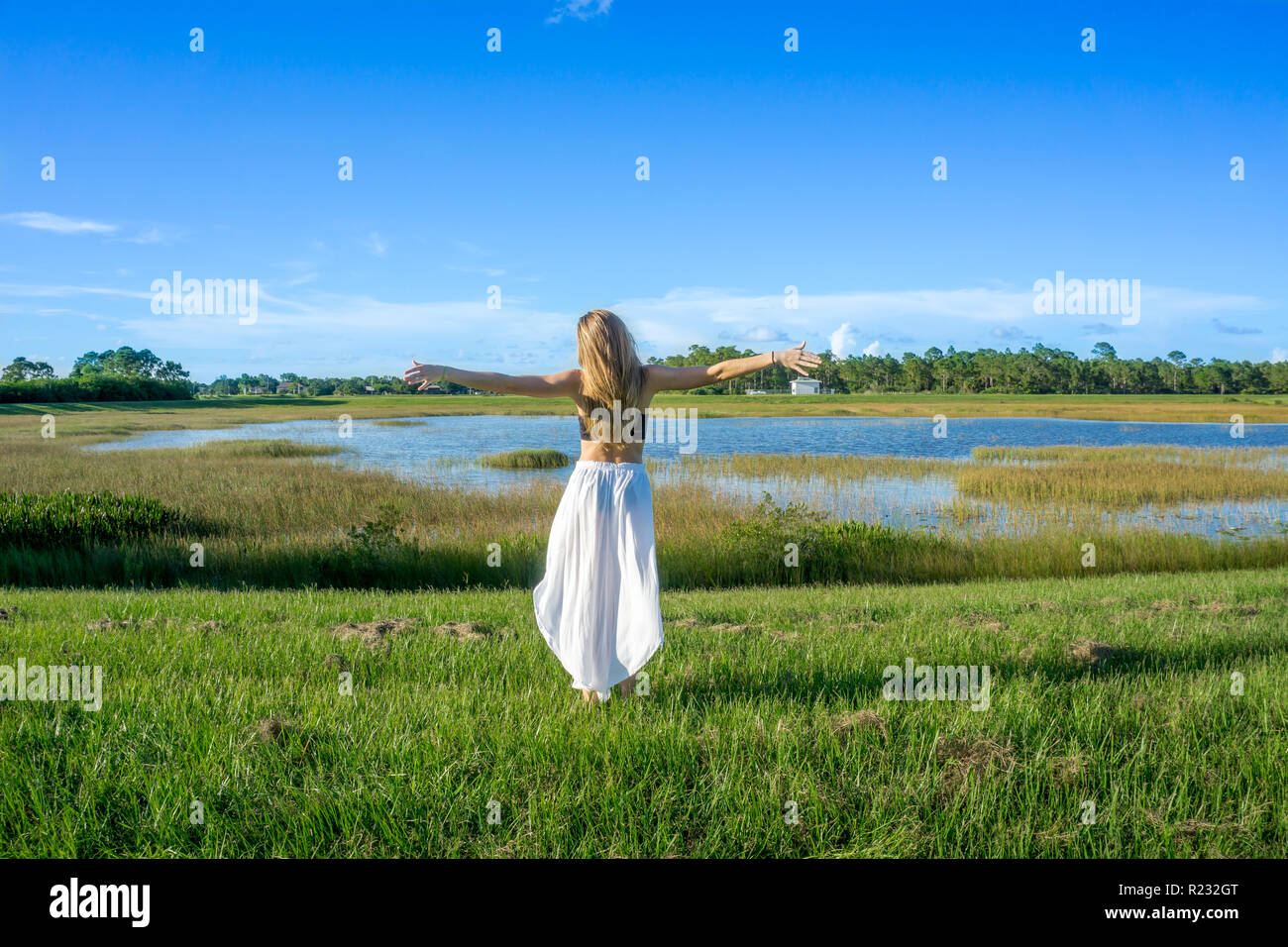 Normale blonde junge Frau mit langen Haaren zurück nach hinten stehend in einem schönen Feld Landschaft draußen mit Hände Arme zum Himmel Stockfoto