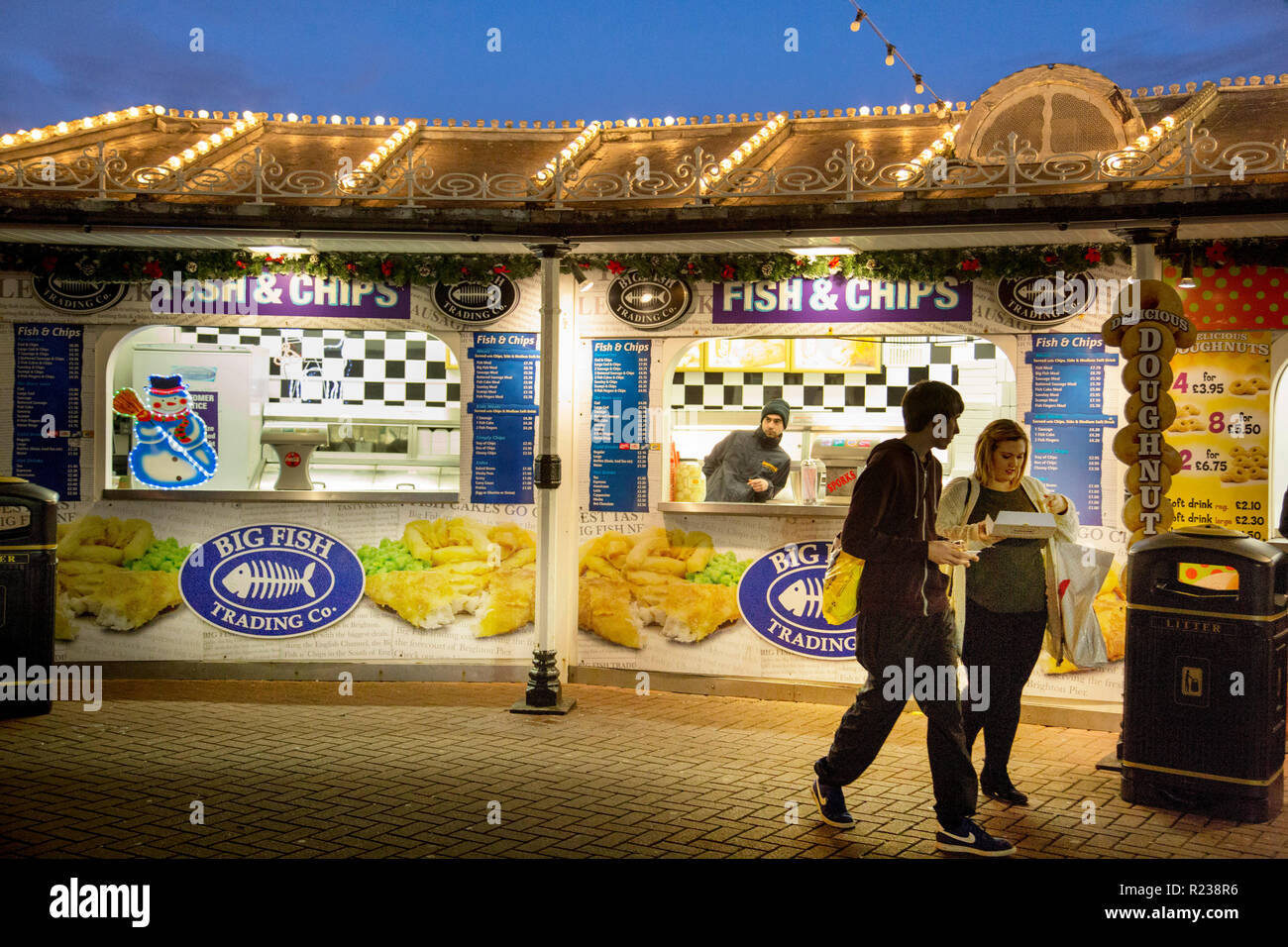 Auf Fisch-und-Chip auf Brighton Pier ein junger Mann und eine Frau Essen kaufen. Overhead, das Dach hat Zeilen von Glühbirnen unter einem Abendhimmel. Stockfoto