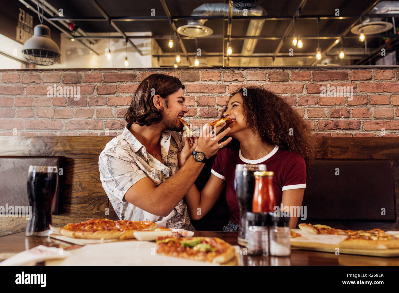 Junger Mann und Frau an Fast Food Restaurant füttern sich gegenseitig mit Pizza sitzen. Paar beim essen Pizza im Cafe. Stockfoto