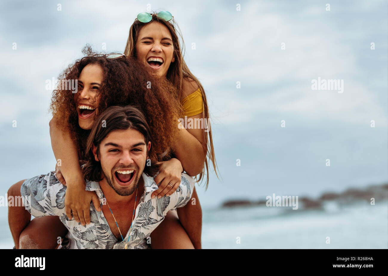 Glückliche junge Mann, der seine Frauen auf seinem Rücken am Strand. Freunde am Meer huckepack. Stockfoto