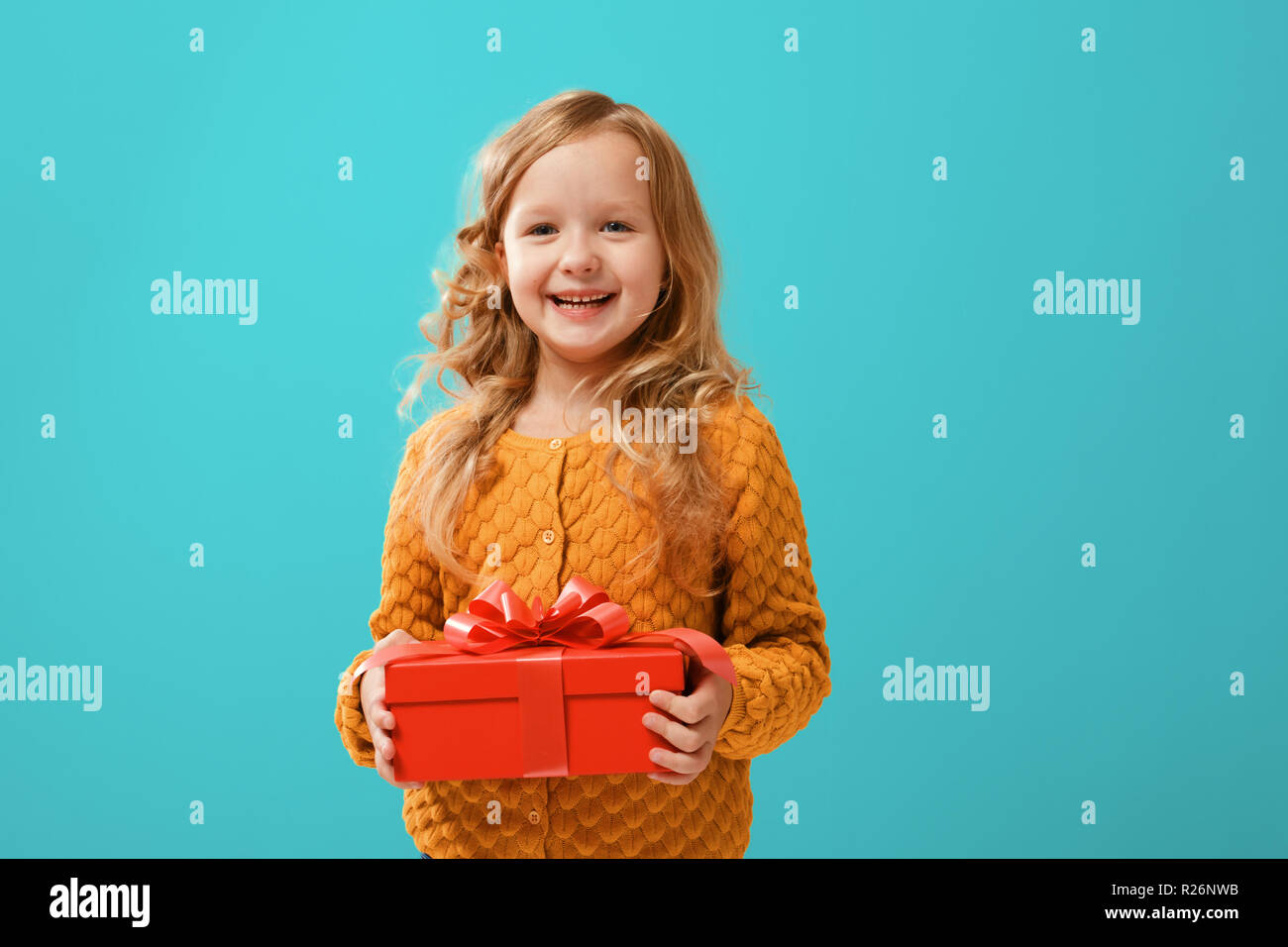 Glückliches Kind Mädchen in einem warmen Senf in Form gestrickter Pullover mit einem roten Kästchen mit einem Geschenk auf einem cyan Hintergrund. Das Konzept der Feier, geben und r Stockfoto