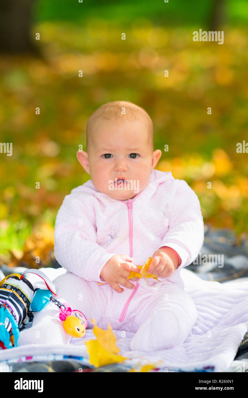 Chubby kleines Mädchen sitzt auf einem Teppich in einem Park von bunten gelben Blätter im Herbst spielt mit ihrem Spielzeug umgeben Stockfoto