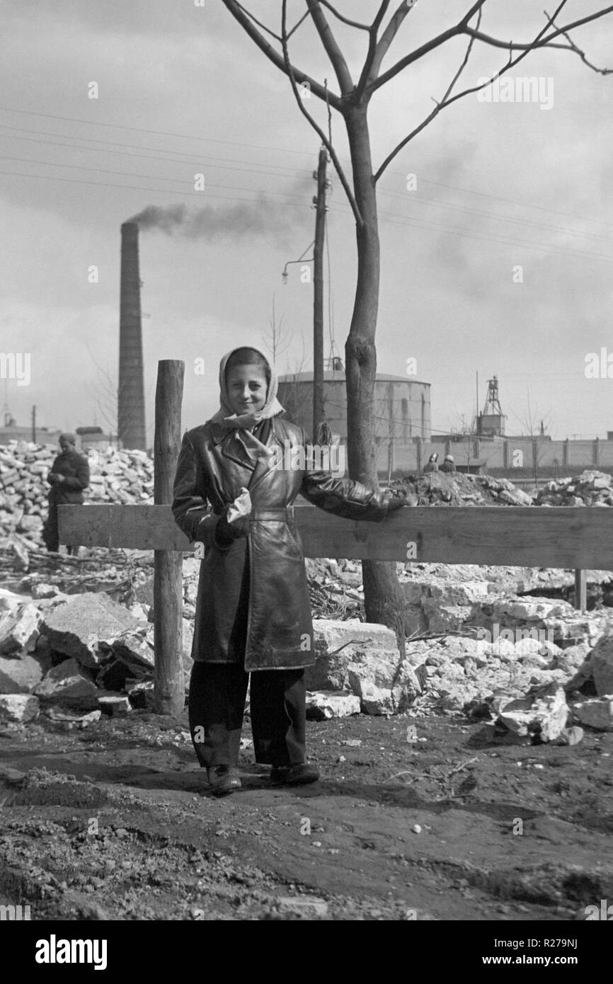 Junge Frau in einem Leder Mantel bekleidet steht in einem trostlosen industriellen urbanen Landschaft mit aufstoßen Schornstein hinter 1950 Ungarn Stockfoto