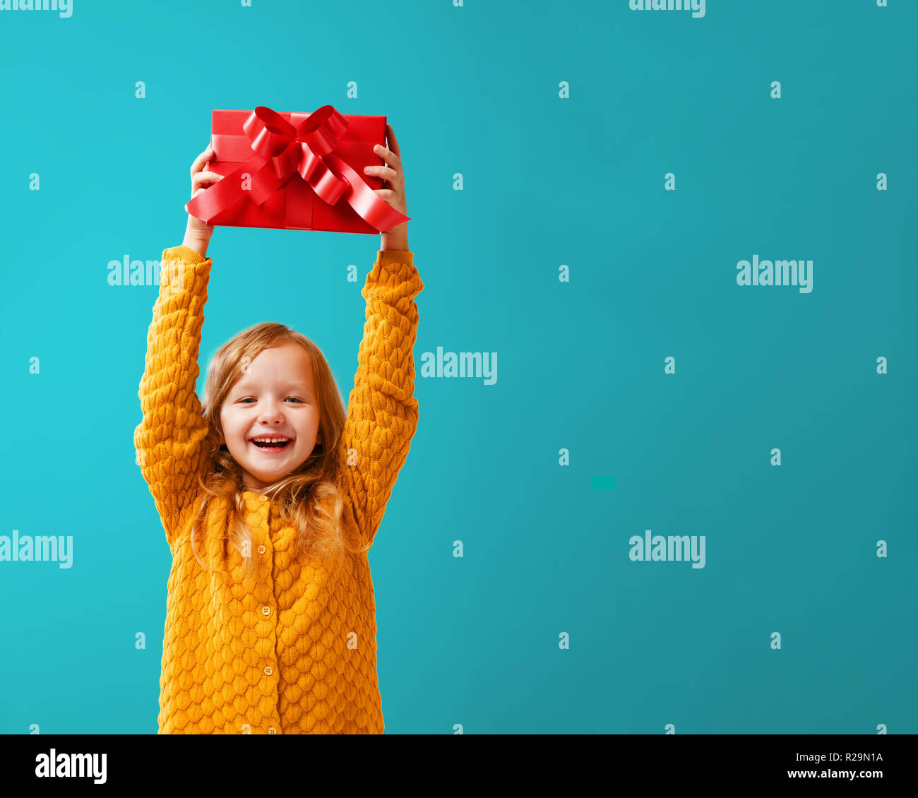 Porträt eines Mädchens in einem warmen Senf in Form gestrickter Pullover auf einem cyan Hintergrund. Ein Kind hält einen roten Kasten mit einem Geschenk über seinem Kopf. Das Konzept der c Stockfoto