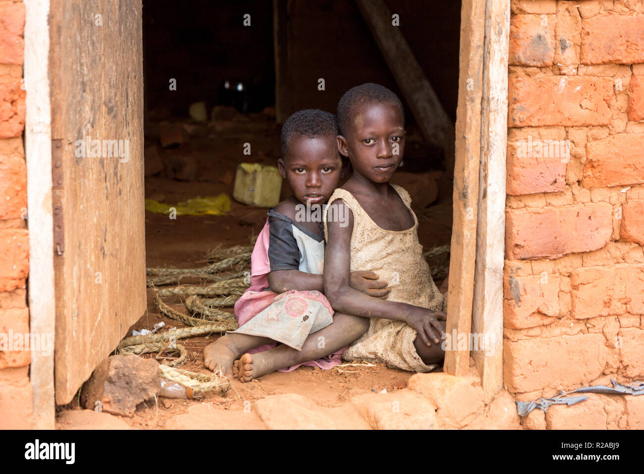 Zwei ugandische Kinder in zerlumpter Kleidung in einer Umarmung auf einem unbefestigten Boden an der Tür eines Hauses sitzen. Stockfoto