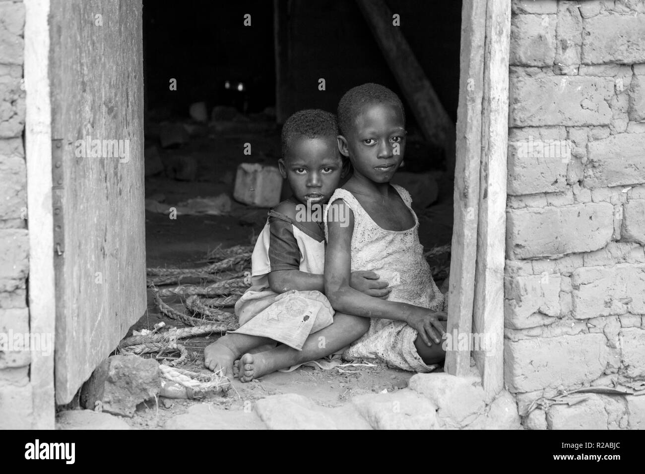 Zwei ugandische Kinder in zerlumpter Kleidung in einer Umarmung auf einem unbefestigten Boden an der Tür eines Hauses sitzen. Stockfoto