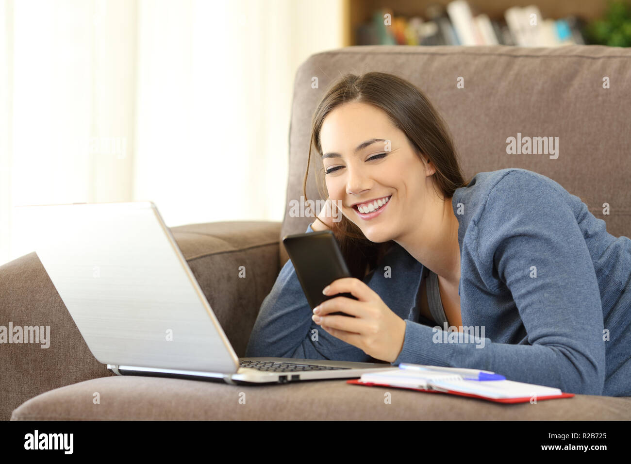Glückliche Frau über ein Telefon und Laptop auf der Couch im Wohnzimmer zu Hause liegen Stockfoto