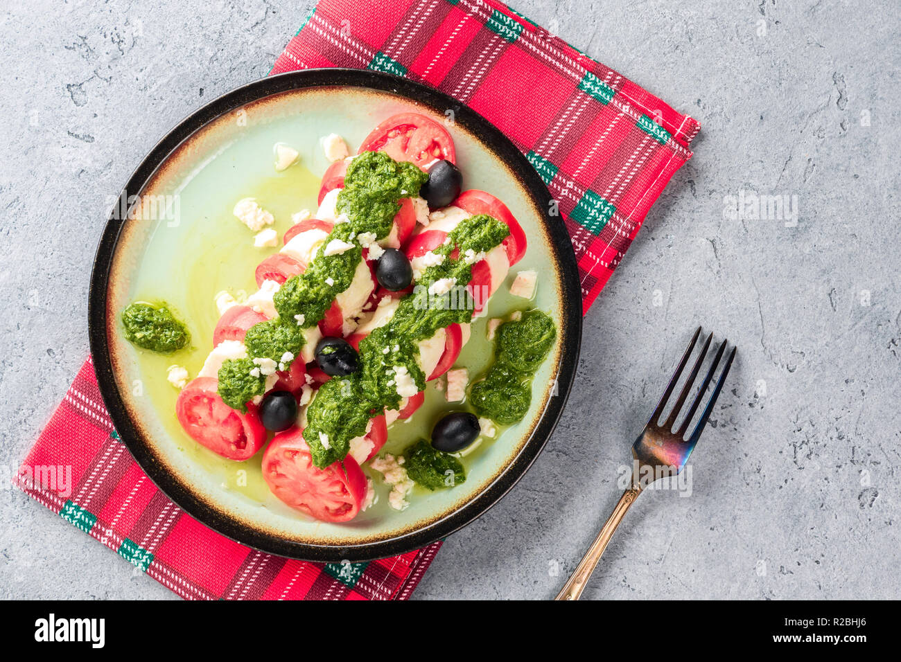 Tomaten und Mozzarella Caprese Salat und Pesto Sauce mit Oliven auf graue Tabelle mit roten überprüft Serviette serviert, Dekanter mit Olivenöl Gabel. Essen Itali Stockfoto