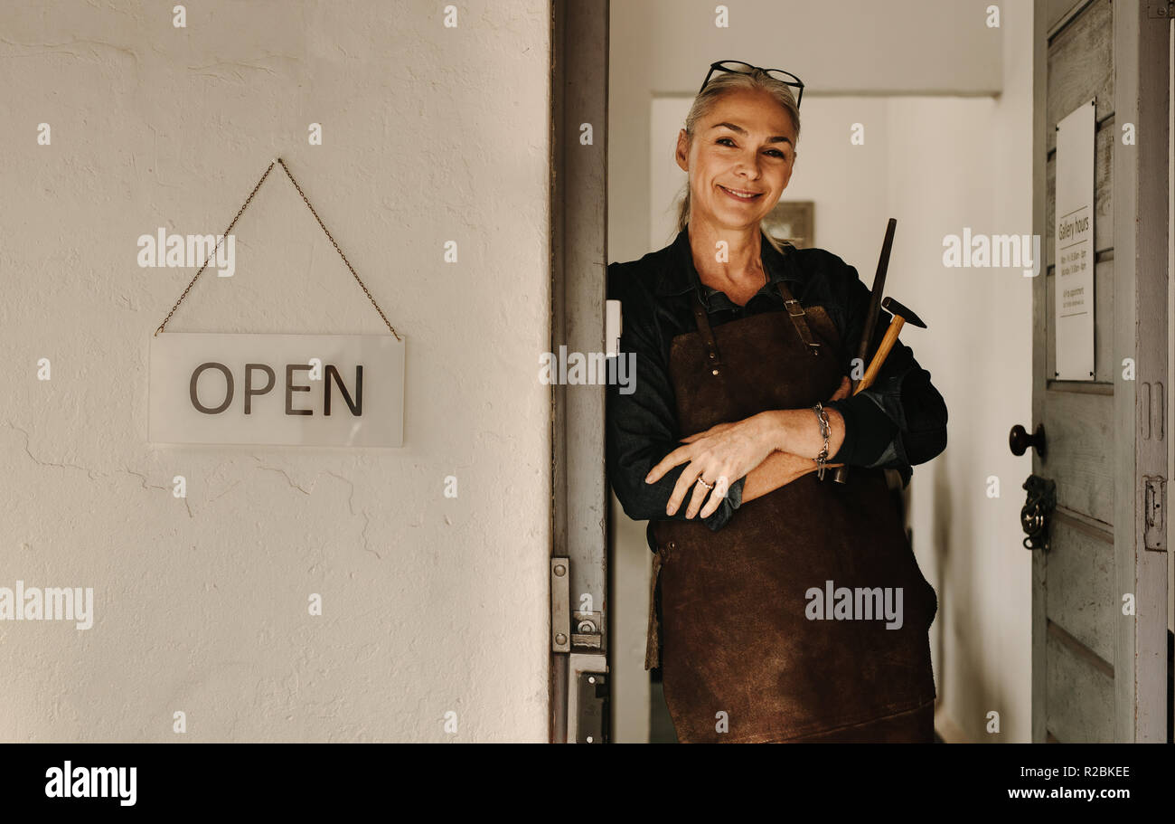 Portrait von lächelnd weibliche Juwelier in Schürze mit Tools von Werkstatt Tür stehen. Senior Juwelier in Schürze können Werkzeuge in der Werkstatt. Stockfoto