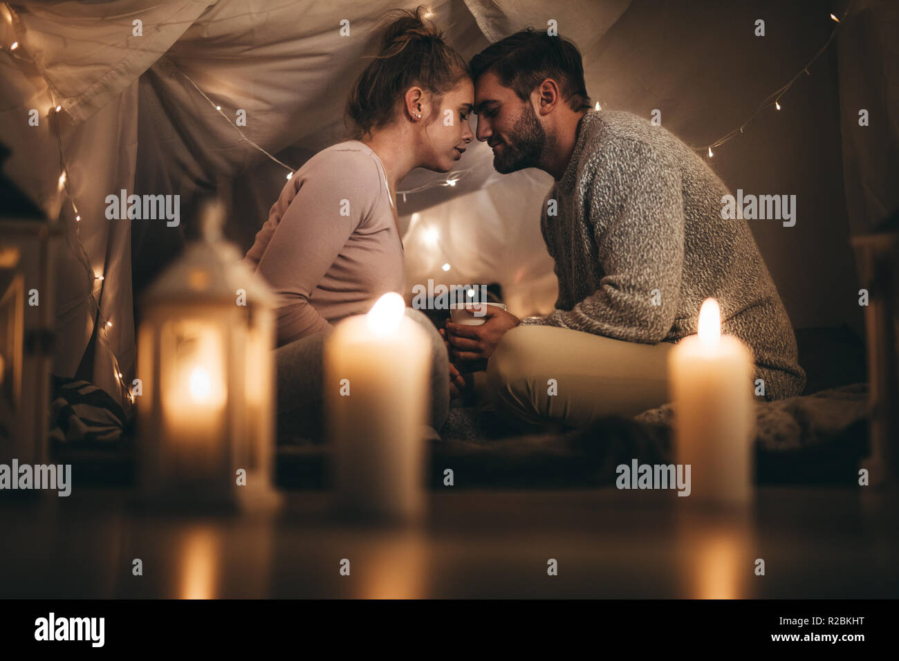 Romantisches Paar sitzt auf dem Bett, das Gesicht berühren ihre Stirn und mit geschlossenen Augen. Mann sitzt auf dem Bett mit seiner Frau hält eine Tasse Kaffee Stockfoto