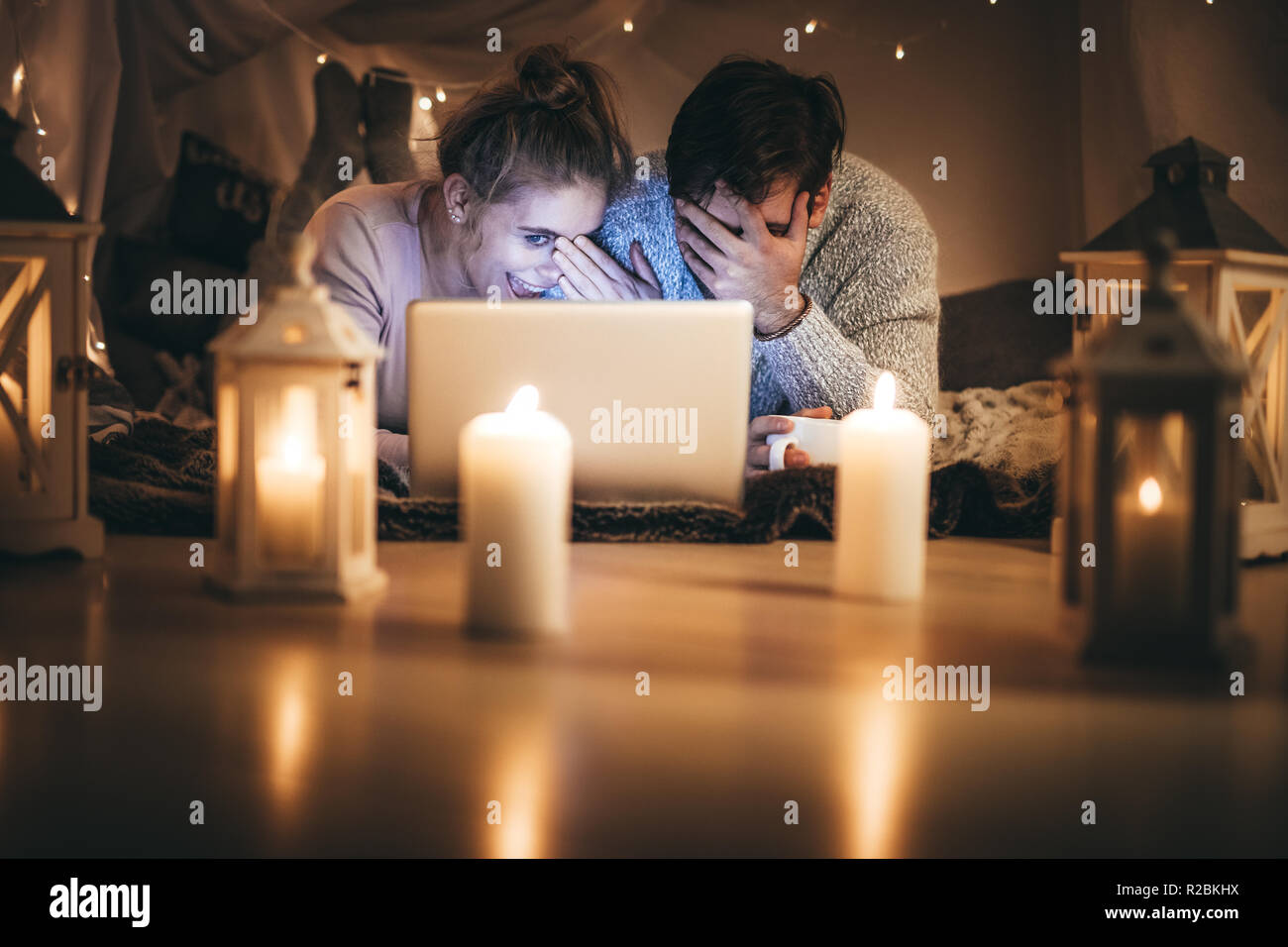 Romantisches Paar liegend auf dem Bett mit einem Laptop in der Nacht in einem Zimmer mit Kerzen geschmückt. Lächeln, Mann und Frau, die ihre Gesichter während an einem l suchen Stockfoto