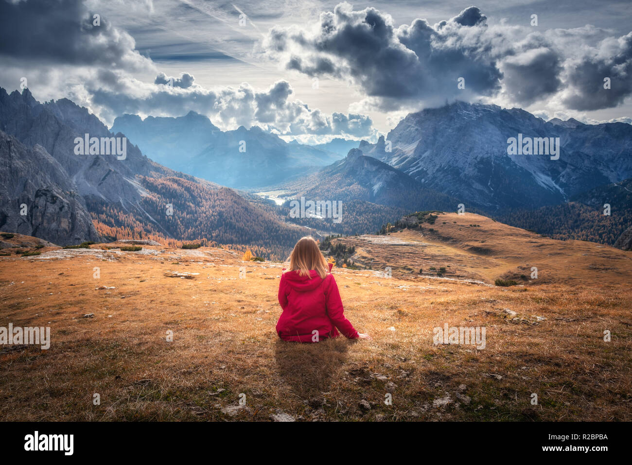 Junge Frau sitzt auf dem Hügel gegen die majestätischen Berge bei Sonnenuntergang im Herbst in den Dolomiten, Italien. Landschaft mit Mädchen, bewölkter Himmel, orange gra Stockfoto
